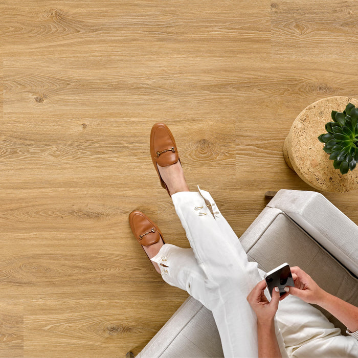 Person sitting on a couch with brown shoes and white pants, holding a phone on an Allure LVP floor.