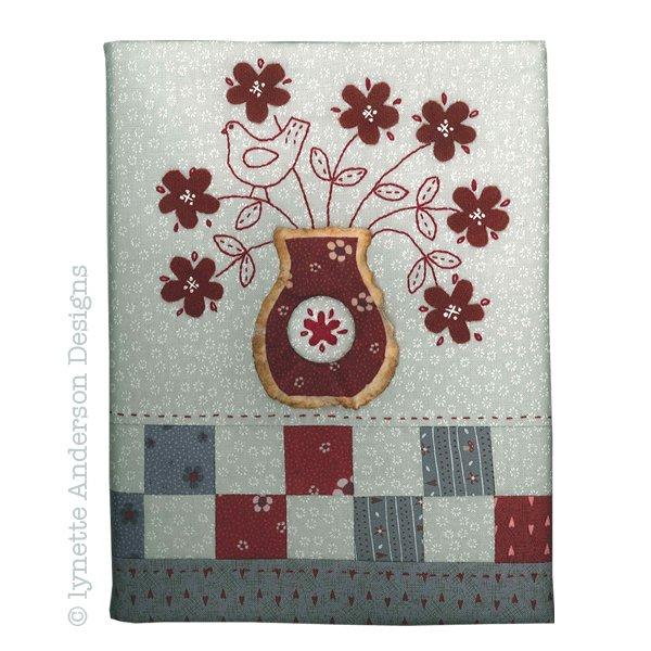 A fabric-covered journal with a floral embroidery design, featuring a bird, flowers, and a vase motif in red, gray, and multicolor on a white background.